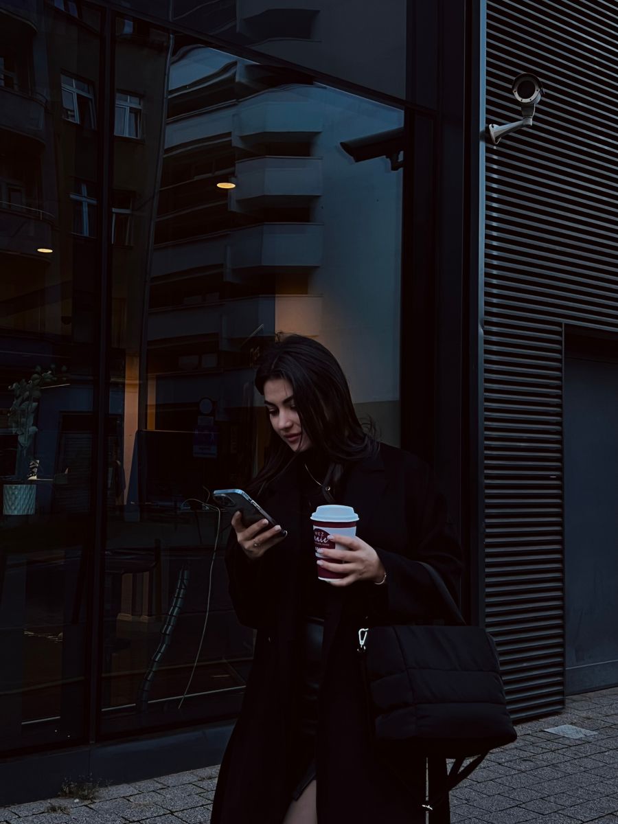 A woman texting on her phone while holding a coffee on the sidewalk